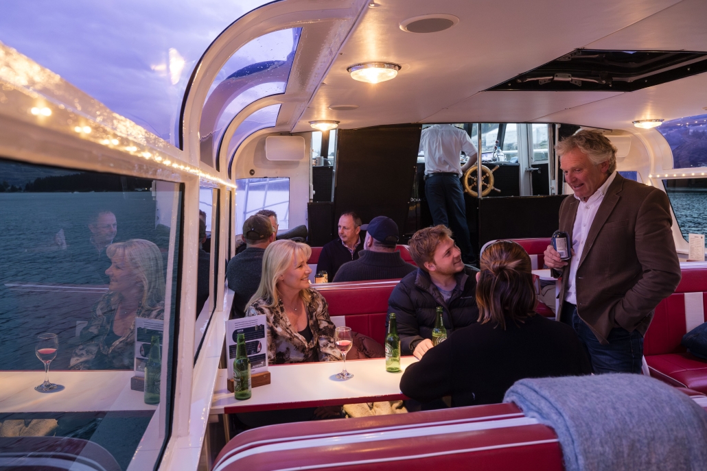 Group of people enjoying drinks and conversation inside a cozy boat lounge at sunset.